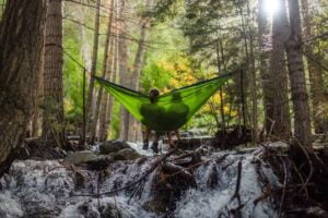 Image of two people in a forested area, resting in a hammock above a stream.