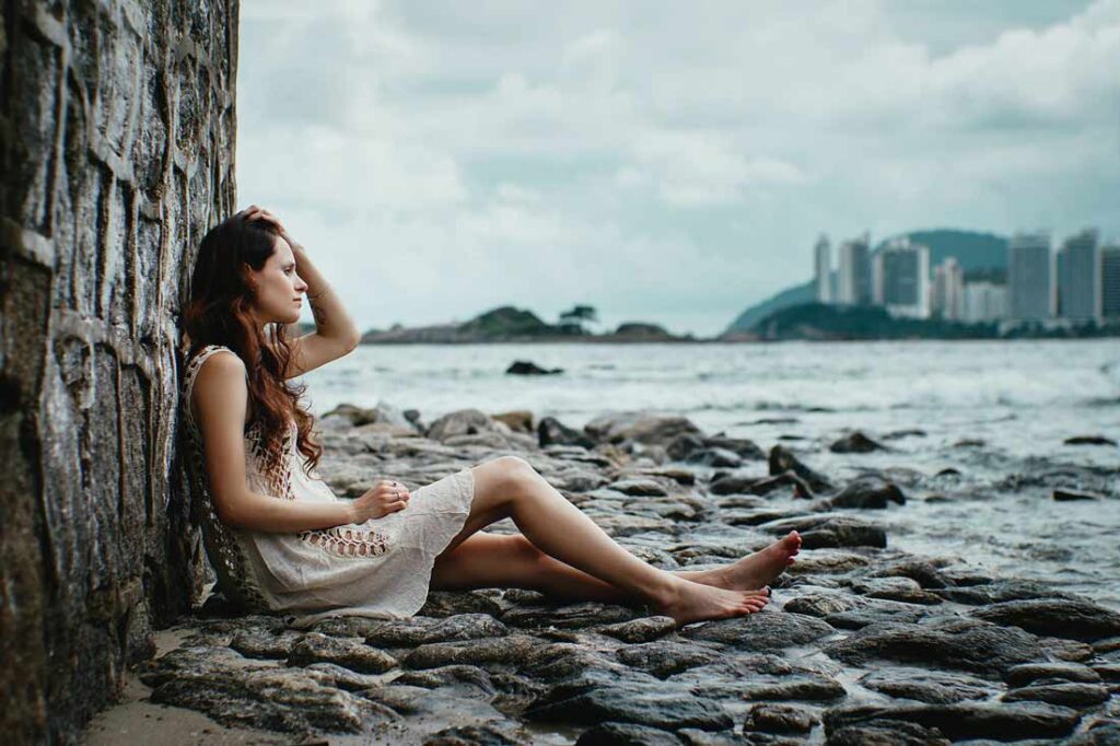 Quitting my 9-5 job was a journey in accepting that the standard framework wasn't the answer for me. It was a journey to accept myself, my desires and my differences. Image of a woman sitting against a rock shelf on a rocky shore on a grey day with a city in the background across a lake.