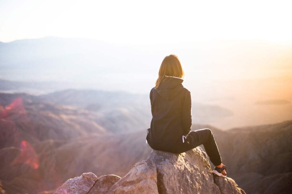 Fear of change doesn't have to hold you back. You can use it to propel you forward by accepting it and taking it in stride. In this article, we'll talk about what you need to do to overcome your fear of change. Image of a woman sitting on a peak looking out over a mountain range at sunrise.