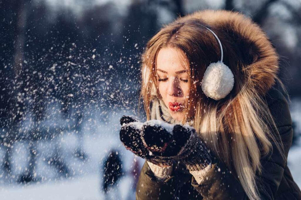 This winter, beat the blues and enjoy the weather. Image of a woman blowing snow from her hands.